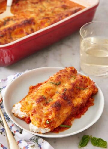 A plate with manicotti with a baking dish and glass of wine in the background.