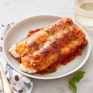 A plate with a serving of manicotti with a glass of wine and a fork beside it.