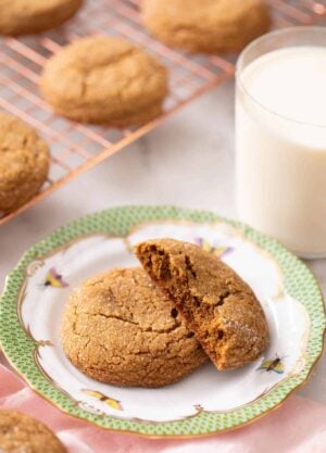 A plate with a molasses cookie with a half cookie slacked on top. A glass of milk in the background and more cookies on a cooling rack.