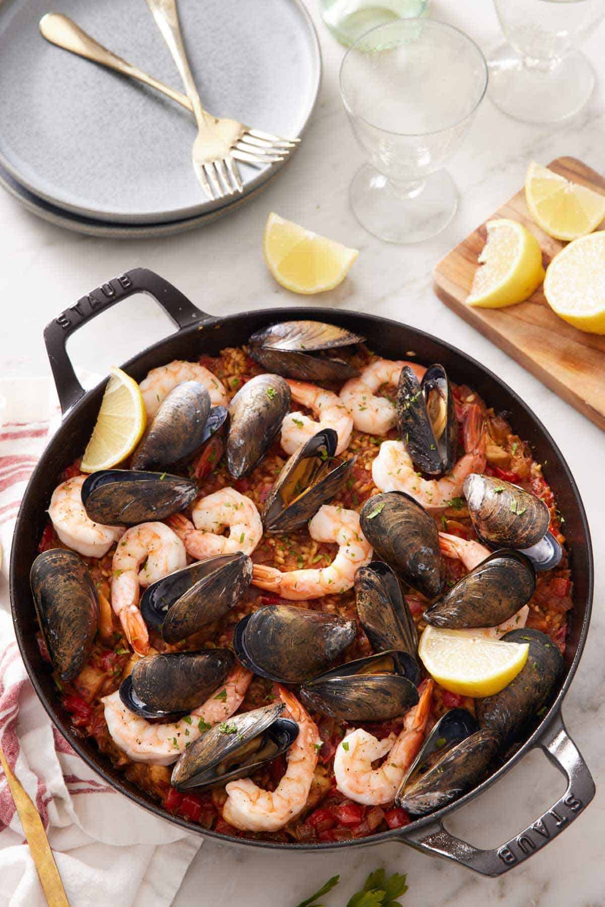 Overhead view of a skillet of paella with a stack of plates, forks, and cut lemon wedges behind it.
