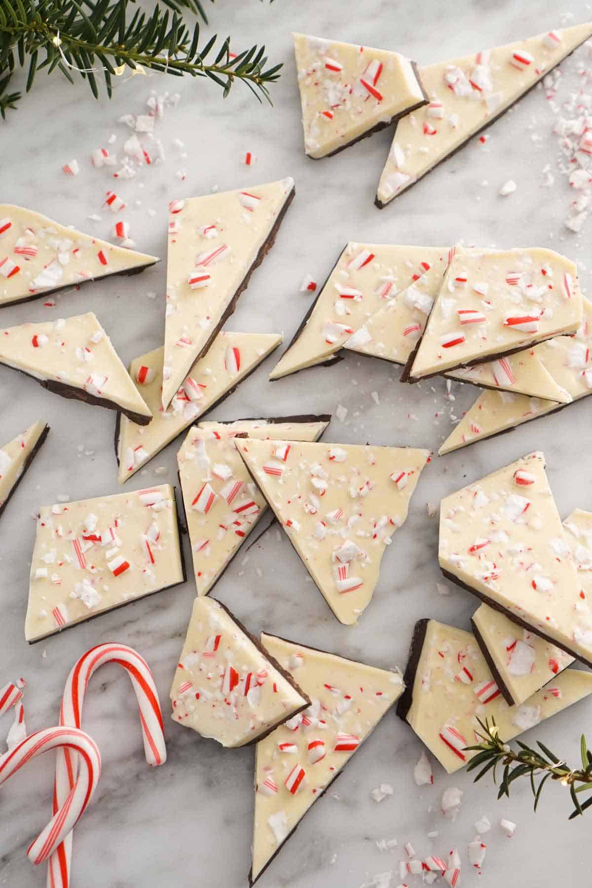 Overhead view of multiple pieces of peppermint bark scattered on a counter surrounded by rosemary and candy canes.