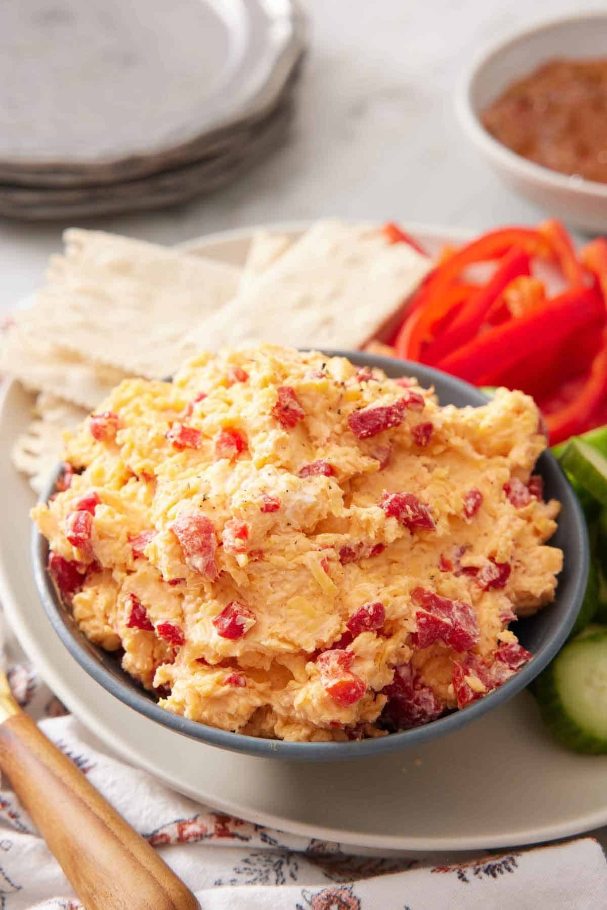 A bowl of pimento cheese along with some crackers, peppers, and cucumbers.
