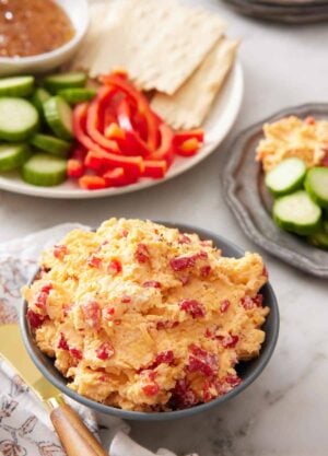 A bowl of pimento cheese with some crackers, peppers, and cucumbers in the background for dipping.