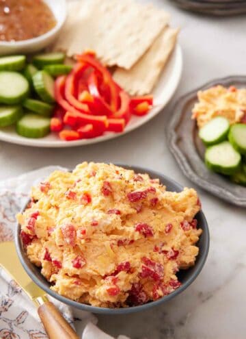 A bowl of pimento cheese with some crackers, peppers, and cucumbers in the background for dipping.