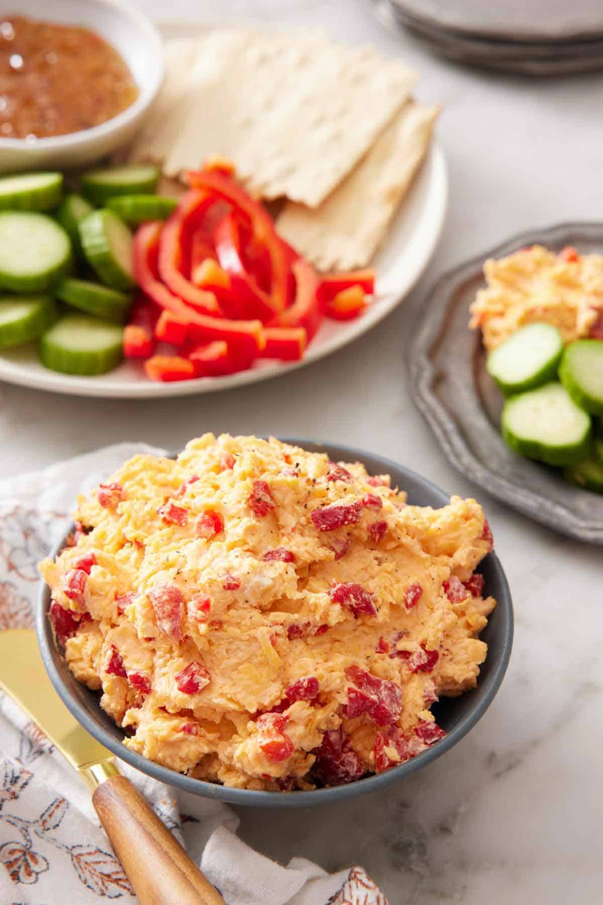 A bowl of pimento cheese with some crackers, peppers, and cucumbers in the background for dipping.