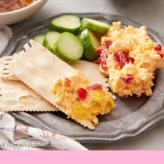 Pinterest graphic of a plate pimento cheese with crackers with some cheese on one and sliced cucumbers beside them.