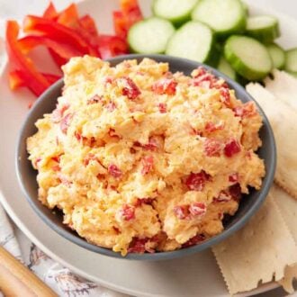 A bowl of pimento cheese with crackers, cucumbers, and bell peppers beside it.