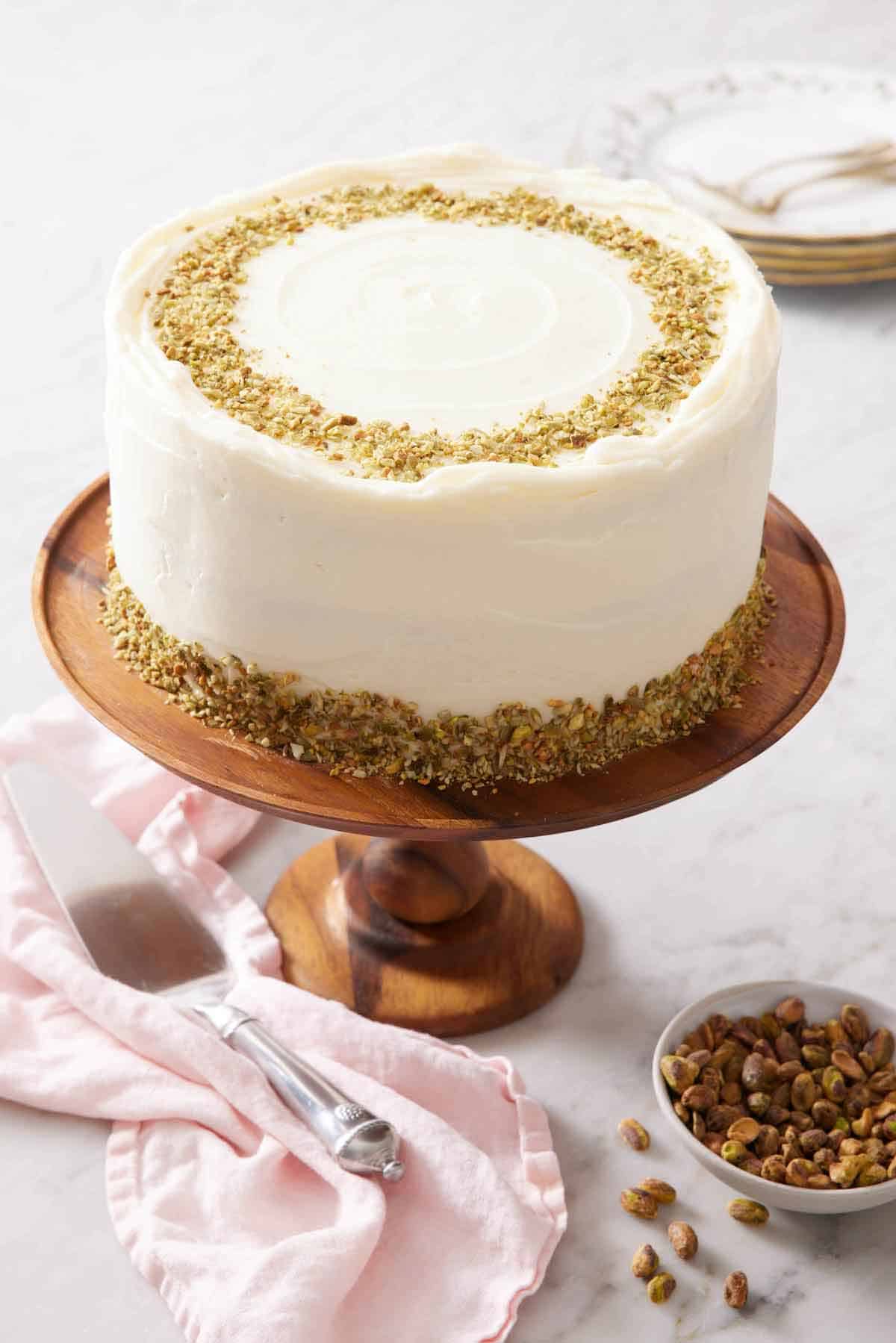 A pistachio cake on a wooden cake stand with a bowl of shelled pistachios on the side.