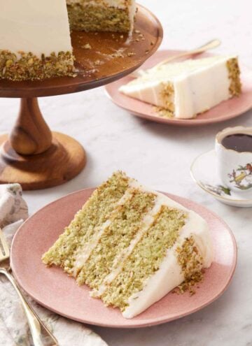 A slice of pistachio cake on a plate with the rest of the cake on a cake stand in the back along with a coffee and second plated slice.