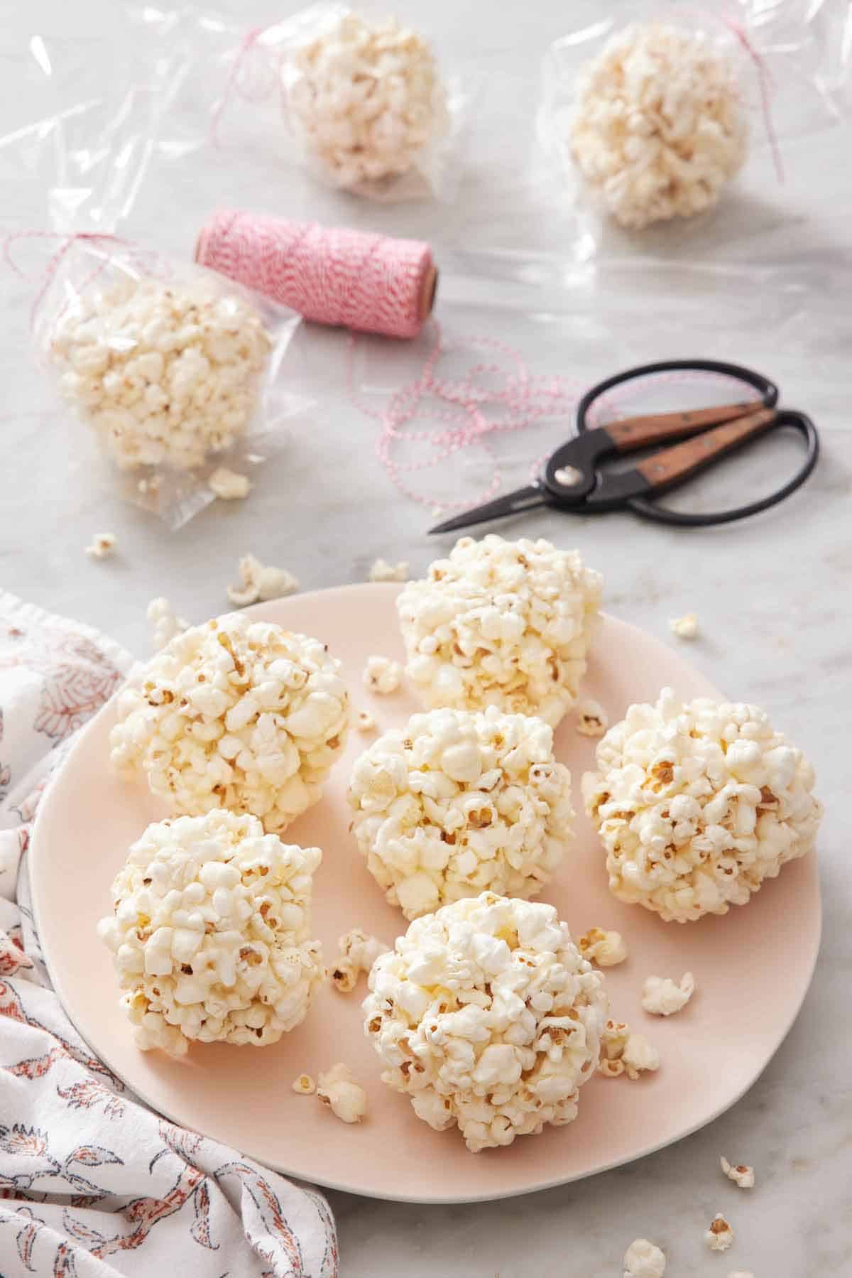 A plate with six popcorn balls. Three packaged popcorn balls in the background with a roll of twine and scissors.