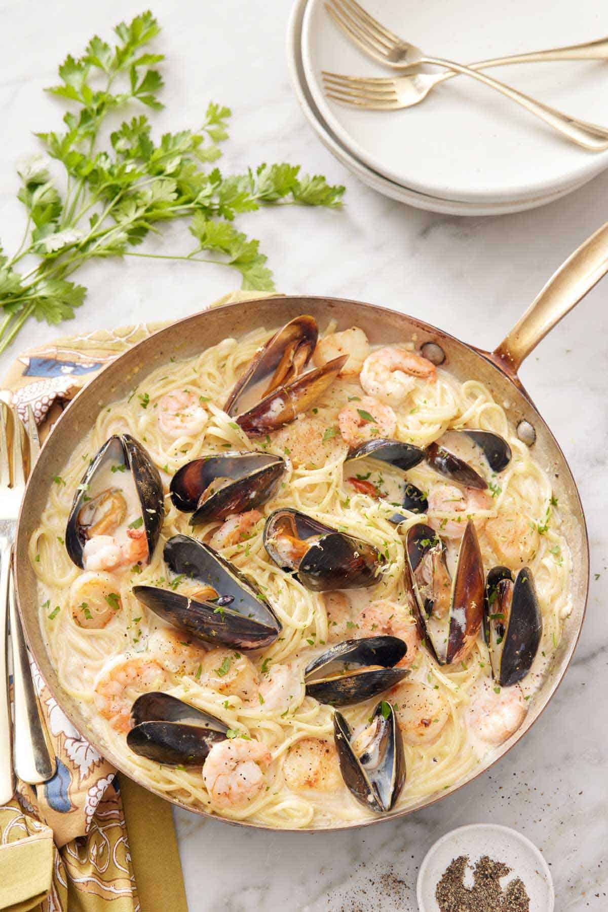 A skillet of seafood pasta with a stack of plates and forks behind it along with parsley.