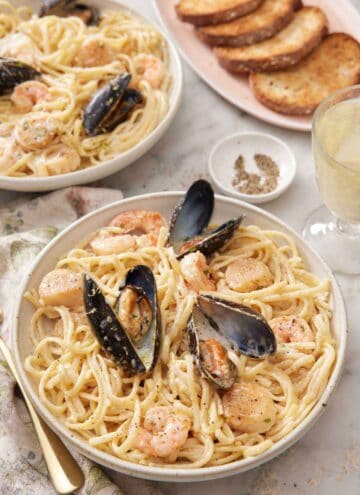 A bowl of seafood pasta with a platter of toasted bread, glass of wine, and another plate in the background.