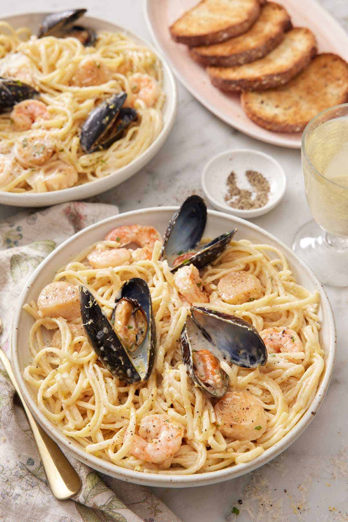 A bowl of seafood pasta with a platter of toasted bread, glass of wine, and another plate in the background.