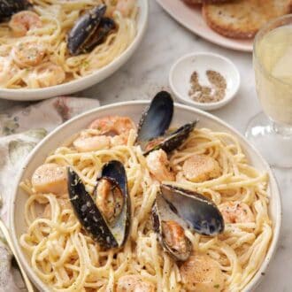 Pinterest graphic of a bowl of seafood pasta with a platter of toasted bread, glass of wine, and another plate in the back.