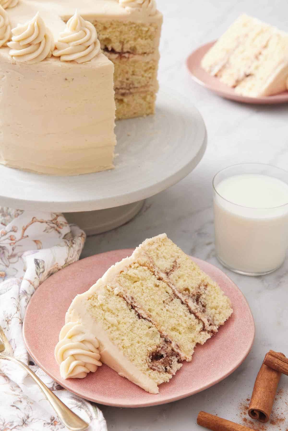 A slice of snickerdoodle cake with a glass of milk and the rest of the cake on a cake stand in the background.