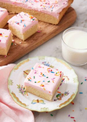 A plate with a sugar cookie bar with a glass of milk and additional sugar cookie bars in the background.