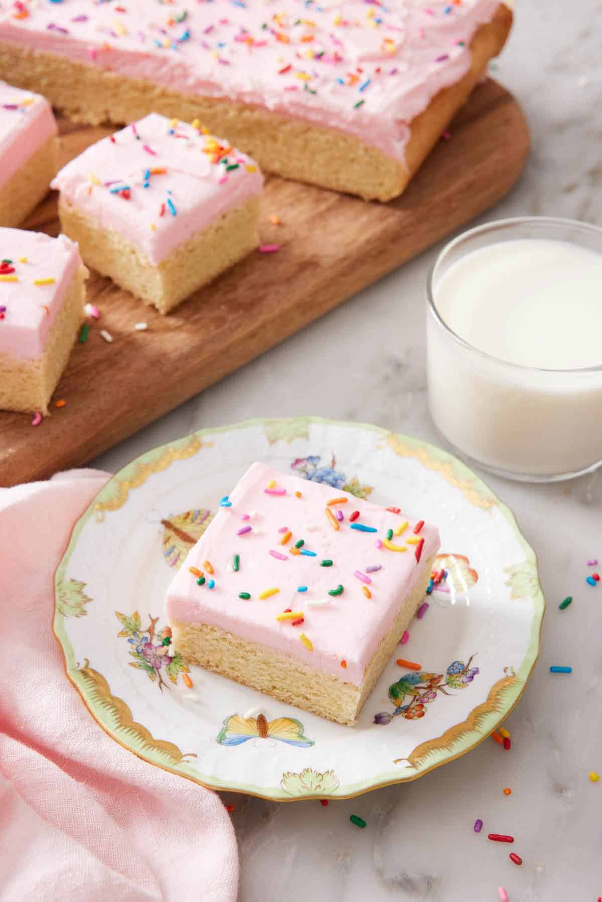 A plate with a sugar cookie bar with a glass of milk and additional sugar cookie bars in the background.