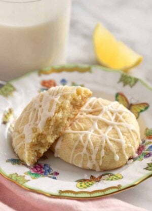 A plate with two lemon cookies, one cut in half. Lemon wedge and glass of milk in the background.