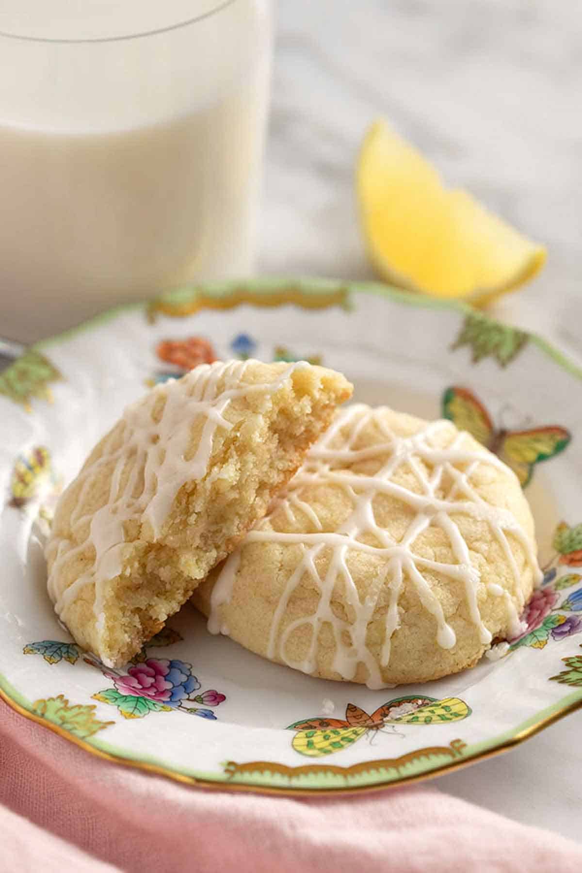 A plate with two lemon cookies, one cut in half. Lemon wedge and glass of milk in the background.