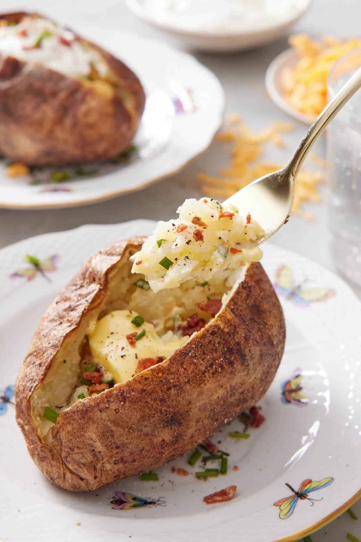 A fork lifting up a bite from a plate with a air fryer baked potato with chives, bacon, and pepper.
