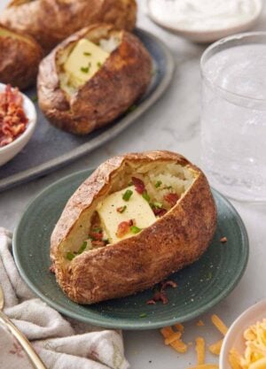 A plate with an air fryer baked potato topped with butter, chives, and crumbled bacon with more potatoes in the background.