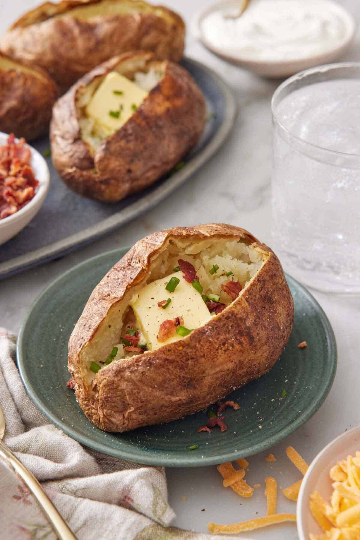 A plate with an air fryer baked potato topped with butter, chives, and crumbled bacon with more potatoes in the background.