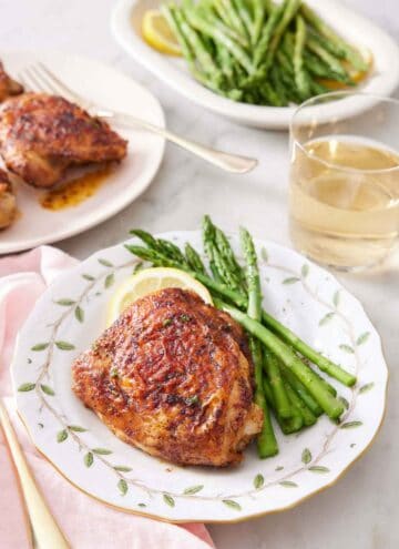 A plate with an air fryer chicken thigh, asparagus, and a lemon slice. A plate of more thighs, a glass of wine, and a platter of asparagus in the background.