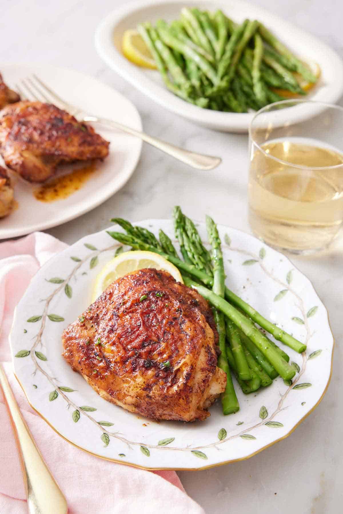 A plate with an air fryer chicken thigh, asparagus, and a lemon slice. A plate of more thighs, a glass of wine, and a platter of asparagus in the background.