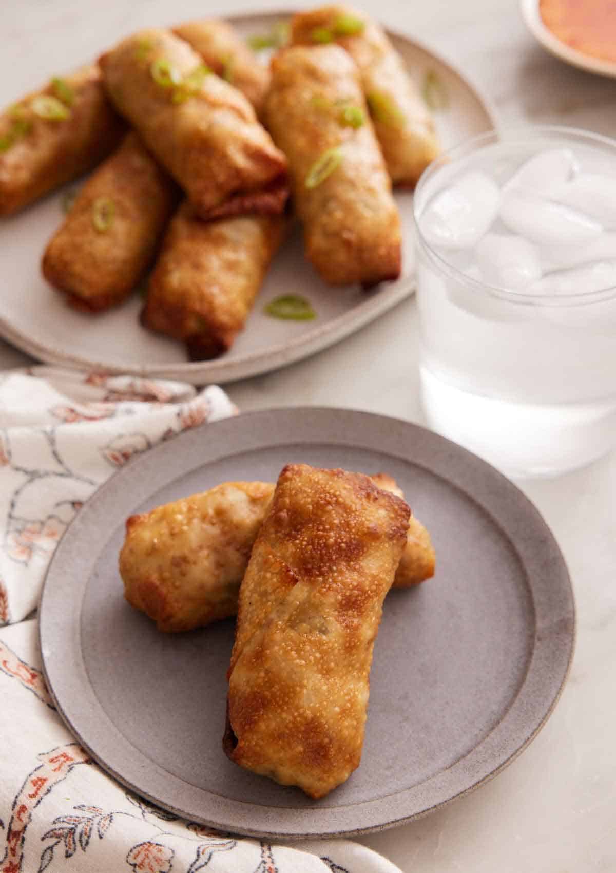 A plate with two air fryer egg rolls with a glass of ice water and platter with more rolls in the background.