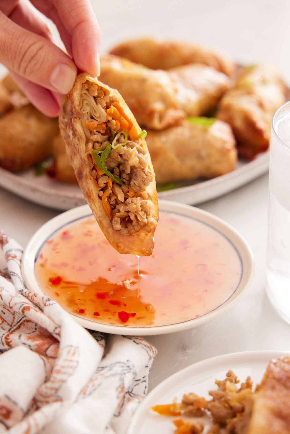 Half an air fryer egg roll dipped in sauce. Platter with more rolls in the background.
