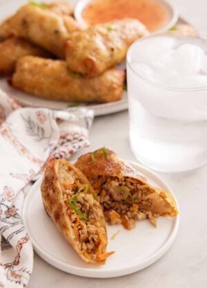 A plate with an air fryer egg roll cut in half. A glass of ice water and platter of more air fryer egg rolls in the background.