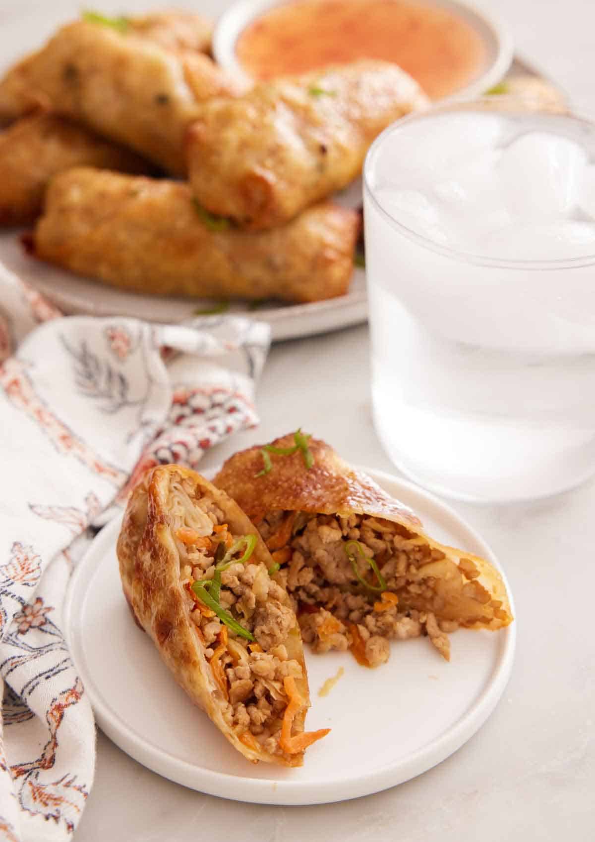 A plate with an air fryer egg roll cut in half. A glass of ice water and platter of more air fryer egg rolls in the background.