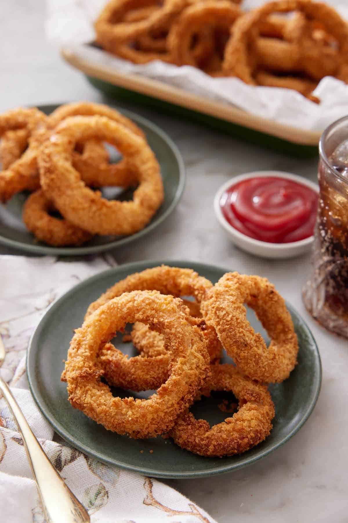 A plate of air fryer onion rings with a second plate and a serving platter with more in the background along with a bowl of ketchup.
