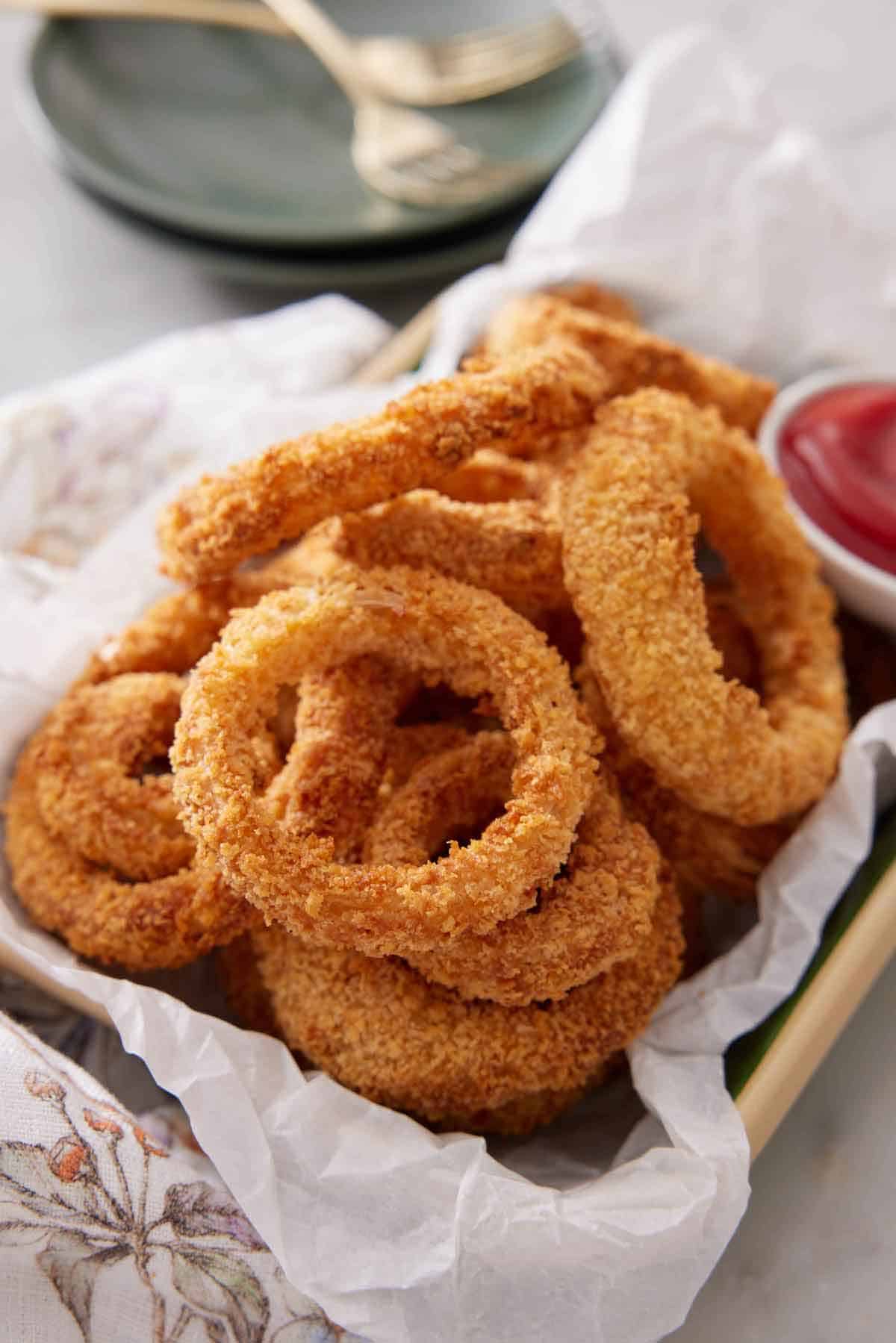 A platter of air fryer onion rings with a small bowl of ketchup with a stack of plate in the background.