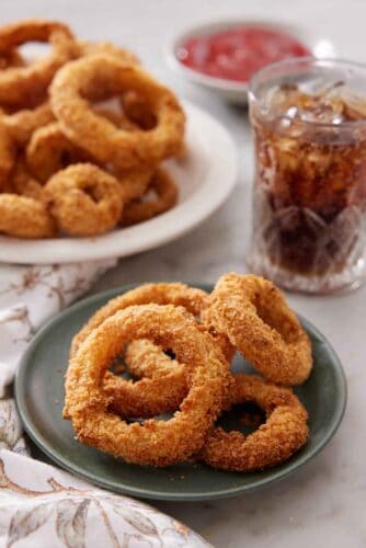 A plate of air fryer onion rings with a platter more in the back along with a glass of iced soda.