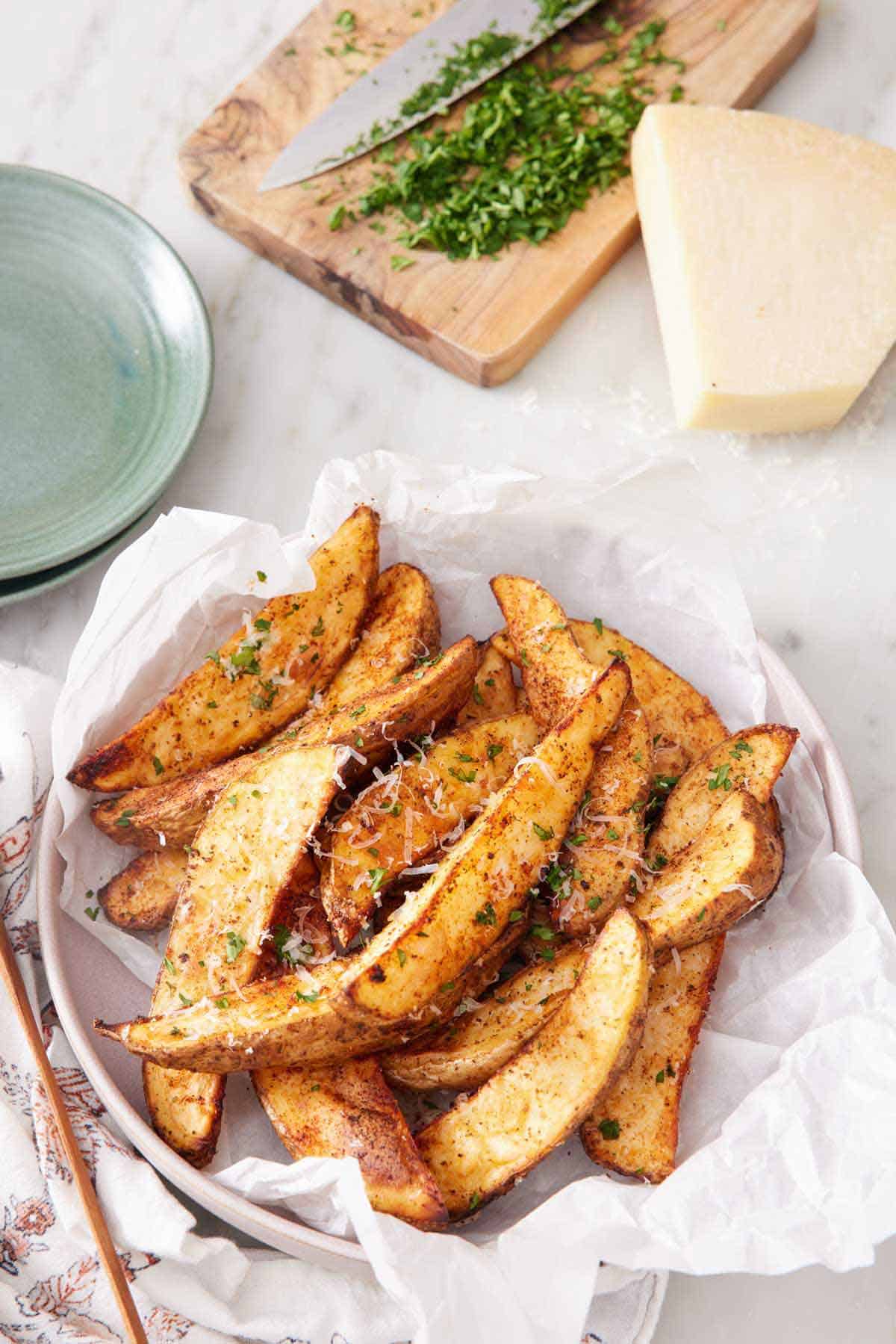 Overhead view of a parchment-lined bowl of air fryer potato wedges topped with parmesan and parsley. A block of parmesan and chopped parsley on a cutting board in the background.