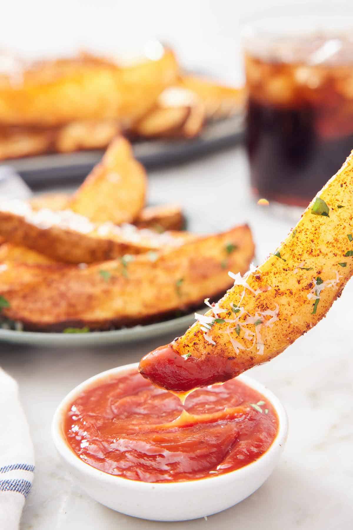 An air fryer potato wedge dipped into a bowl of ketchup. More wedges in the background and a glass of soda.
