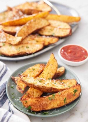 A plate of air fryer potato wedges topped with grated parmesan and parsley with a platter more in the background with a bowl of ketchup.