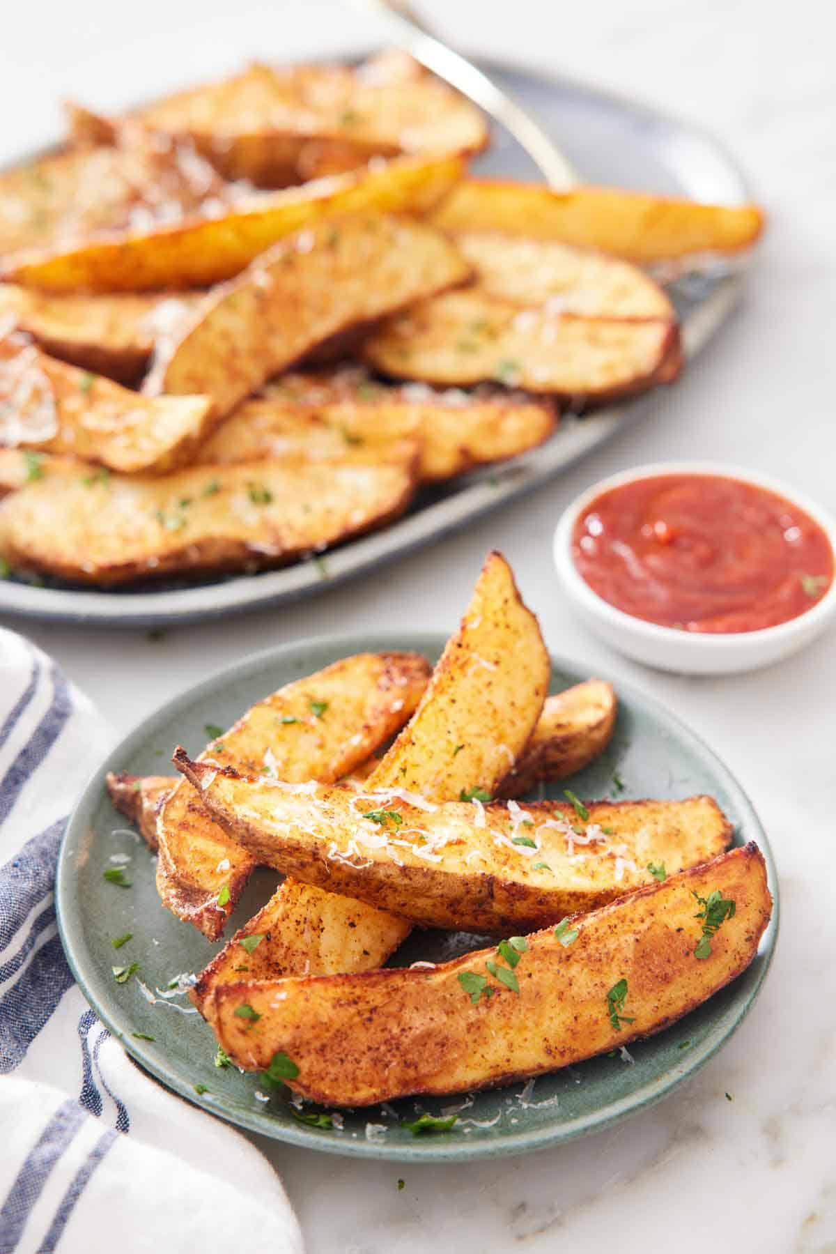 A plate of air fryer potato wedges topped with grated parmesan and parsley with a platter more in the background with a bowl of ketchup.