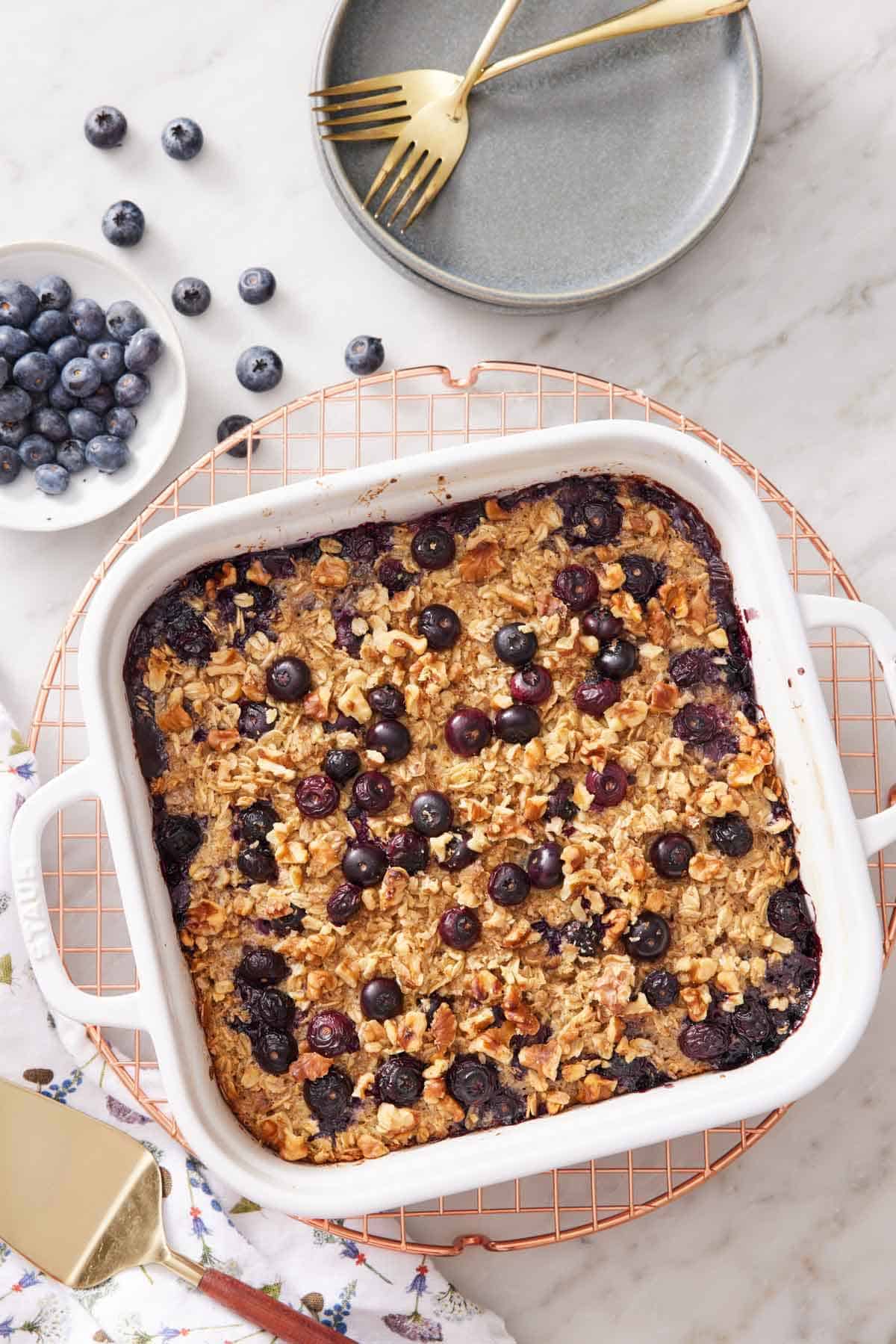 Overhead view of a white baking dish with baked oatmeal on a cooling rack with a bowl of blueberries beside it with some scattered. Forks and plates beside it.