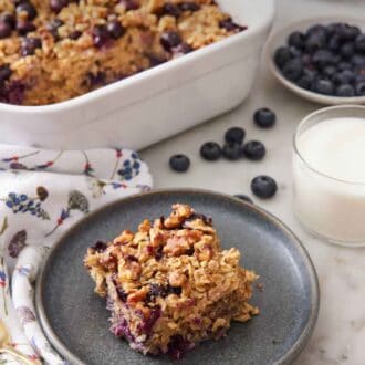 Pinterest graphic of a plate with a square piece of baked oatmeal. Baking dish of the rest of the oatmeal, a bowl of blueberries, and a glass of milk in the back.