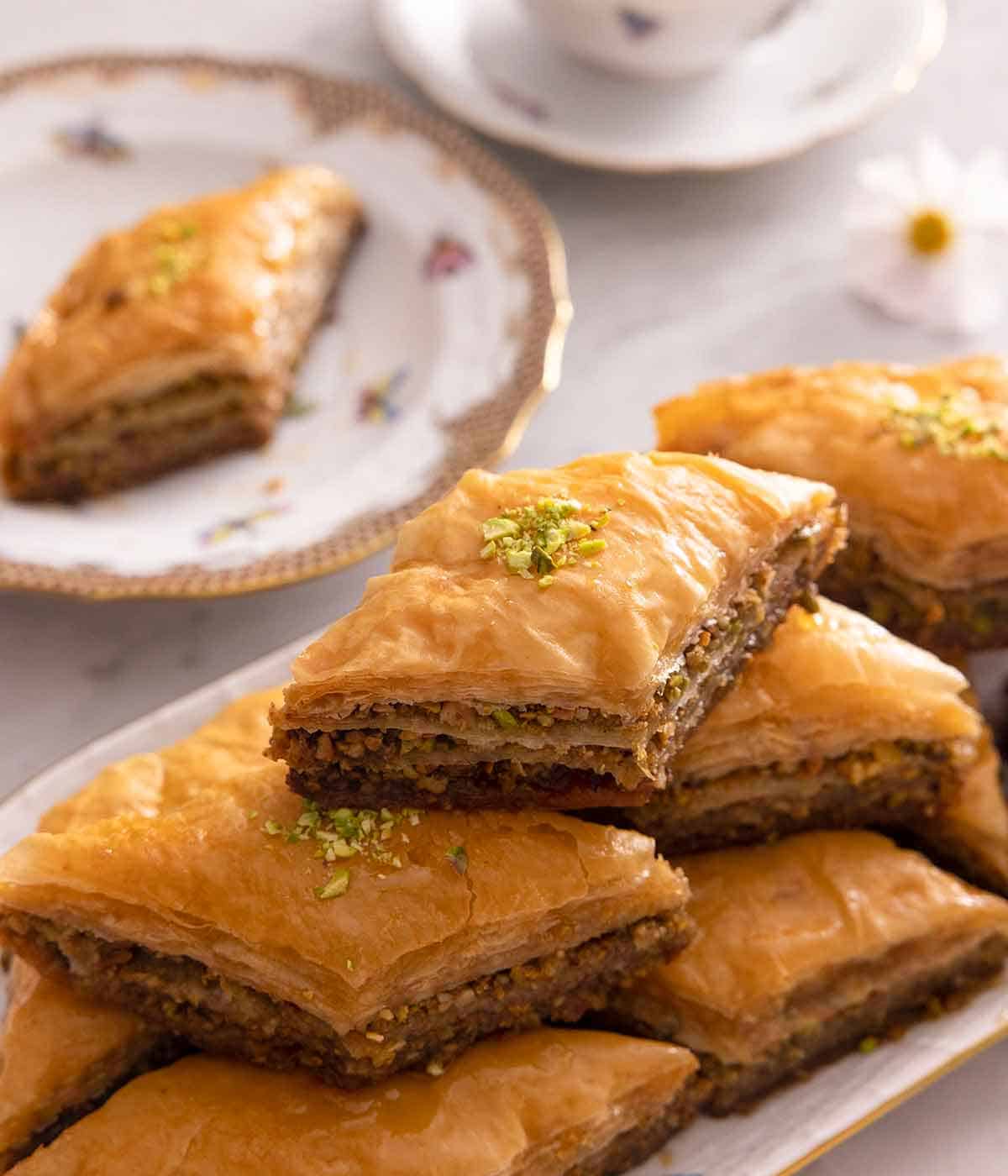 A platter of baklava stacked in three layers with a plate with one baklava in the back.