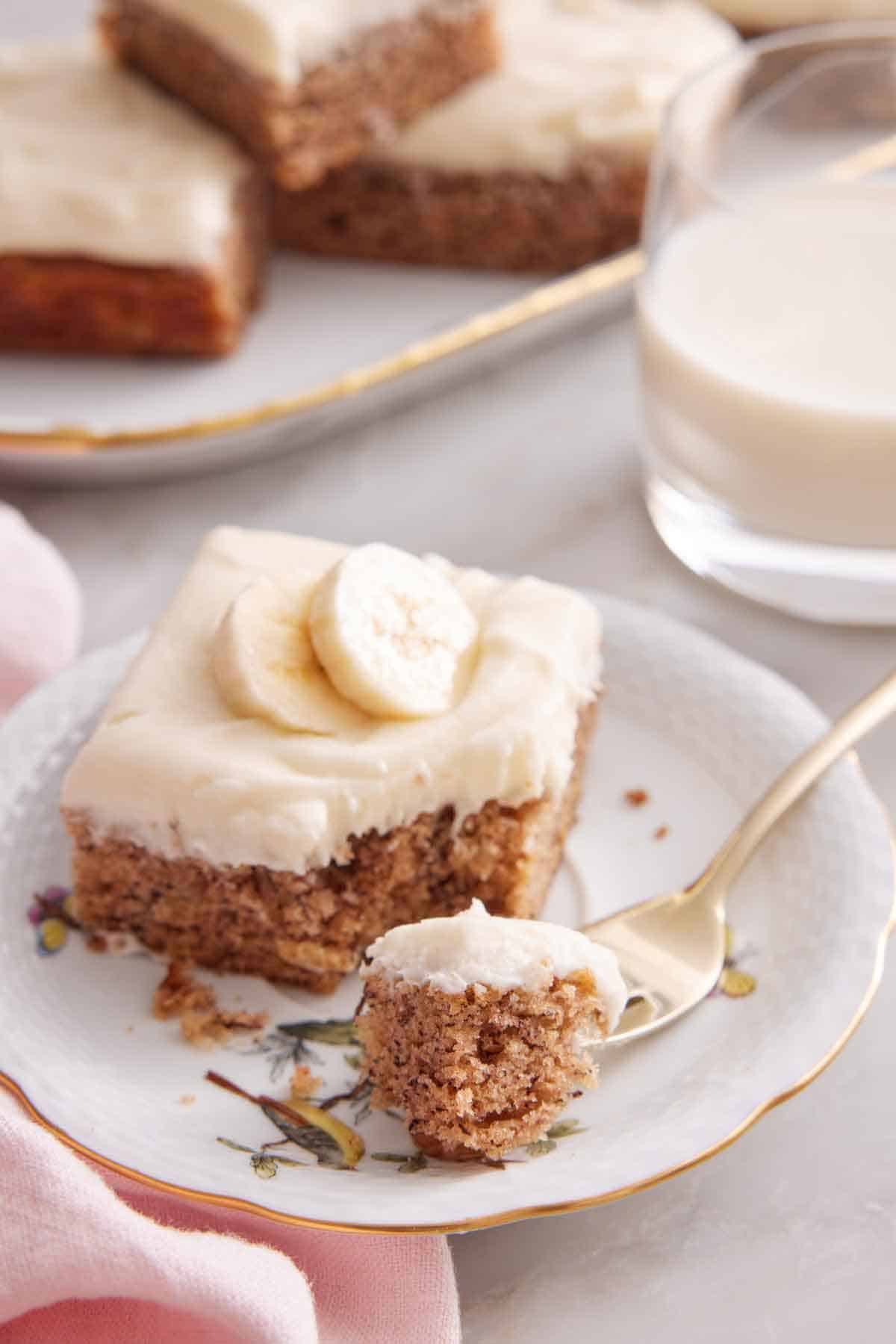 A plate with a slice of banana bar with a corner bite on a fork. Glass of milk and more bars in the back.