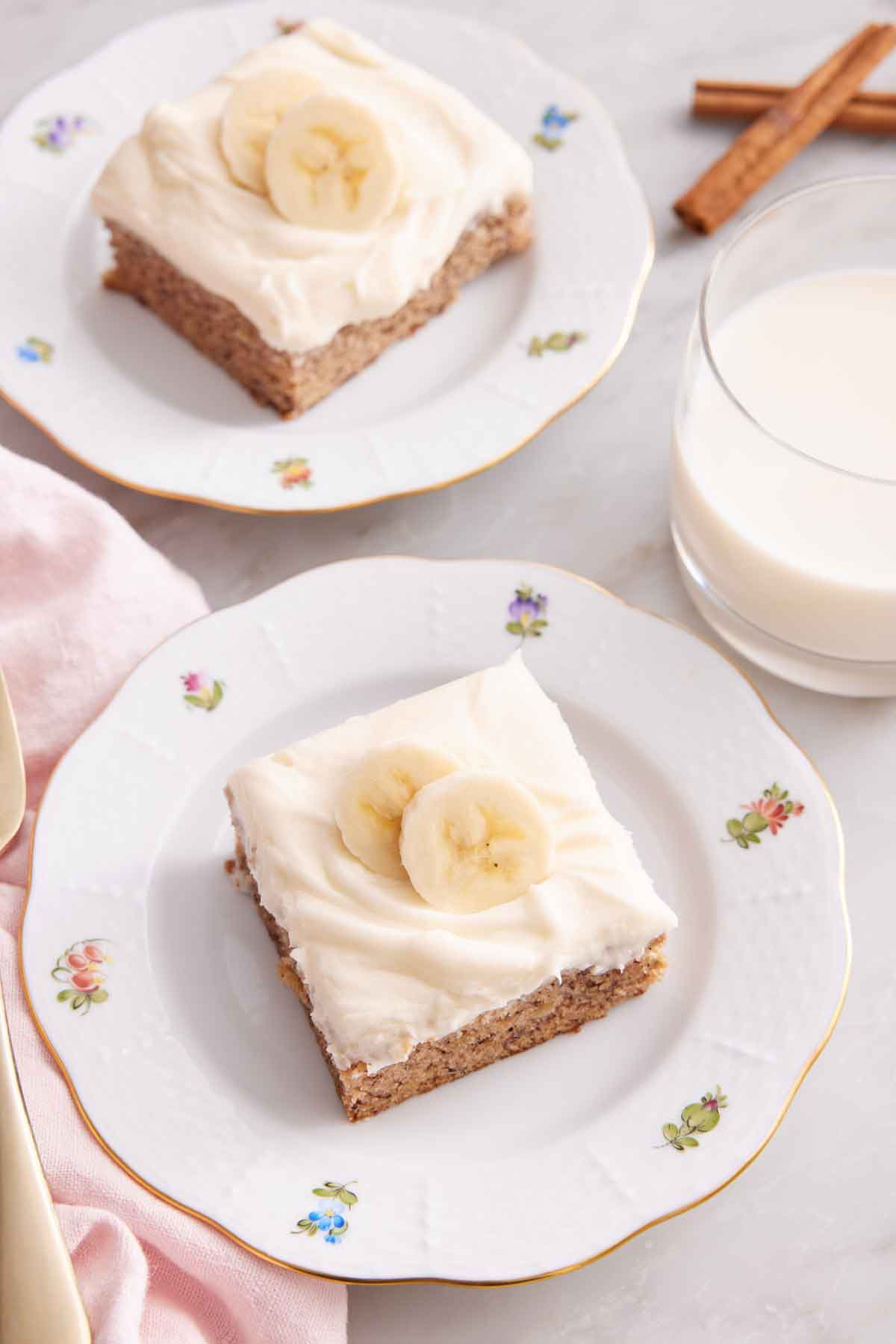 Two plates of banana bars with sliced bananas on top. Glass of milk to the side.