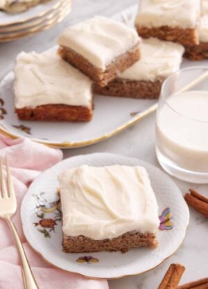 A plate with a slice of banana bar with a glass of milk and platter of more bars in the background.