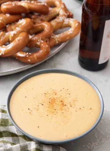 A bowl of beer cheese dip with a platter of pretzels and a bottle of beer in the background.