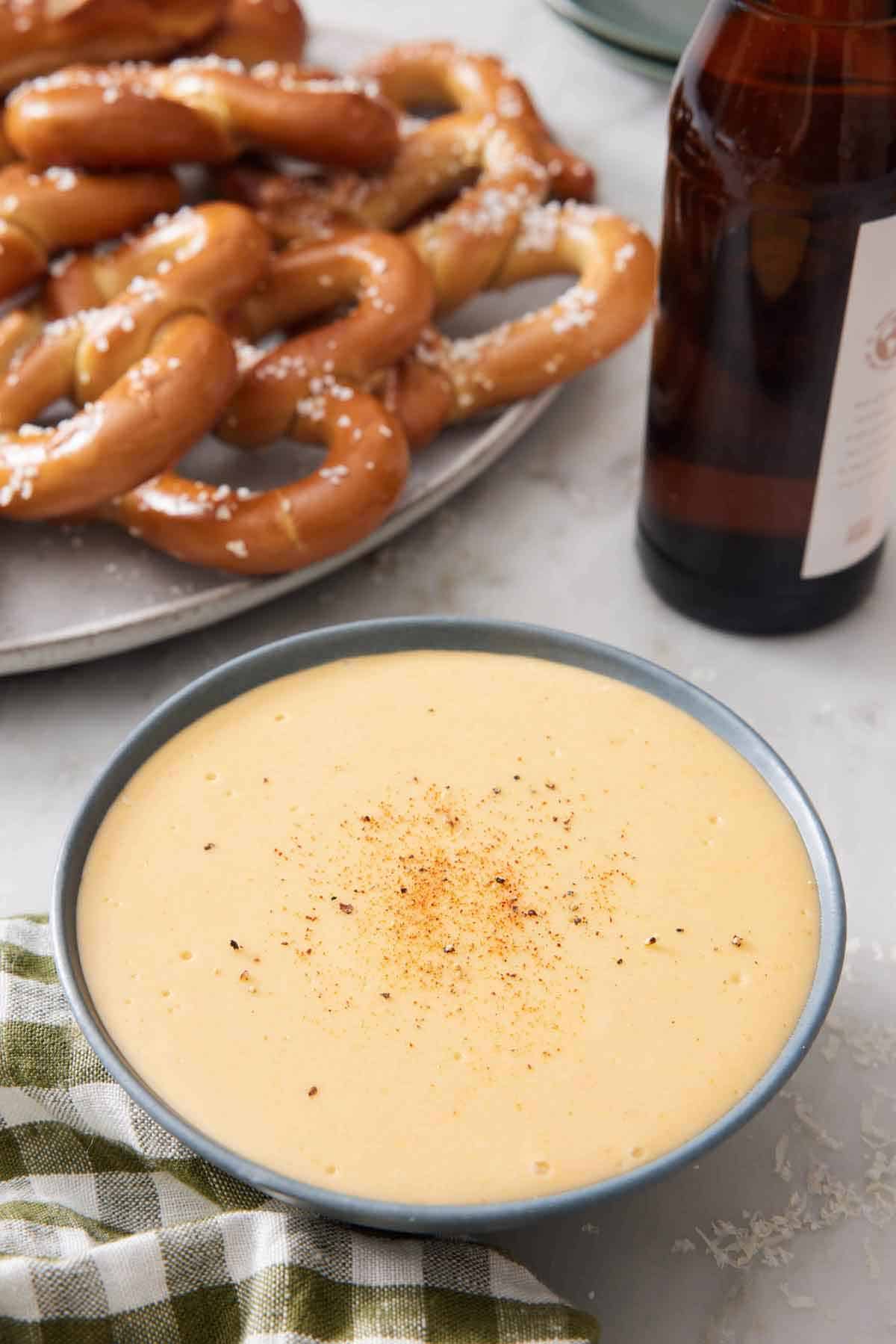 A bowl of beer cheese dip with a platter of pretzels and a bottle of beer in the background.