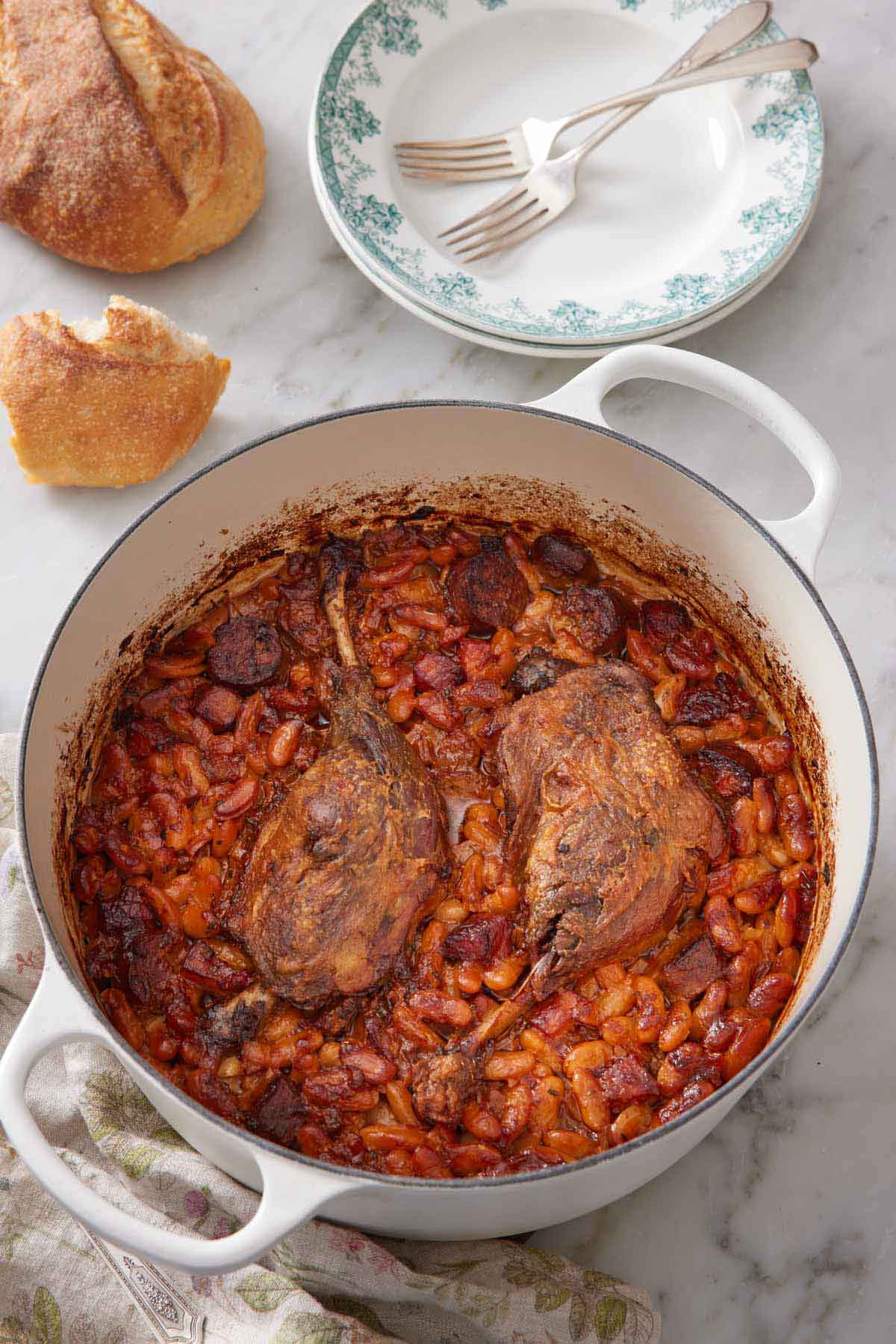 A pot with cassoulet with a couple of plates and forks in the background along with torn bread.