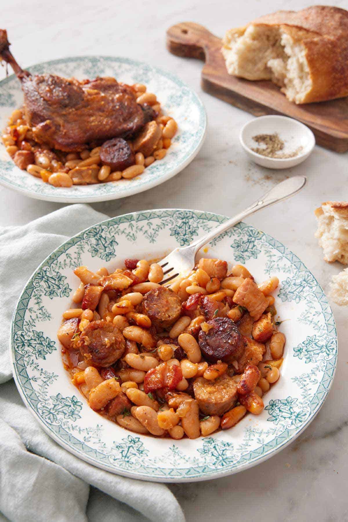 A plate of cassoulet with a fork with a second plate and torn bread in the background.