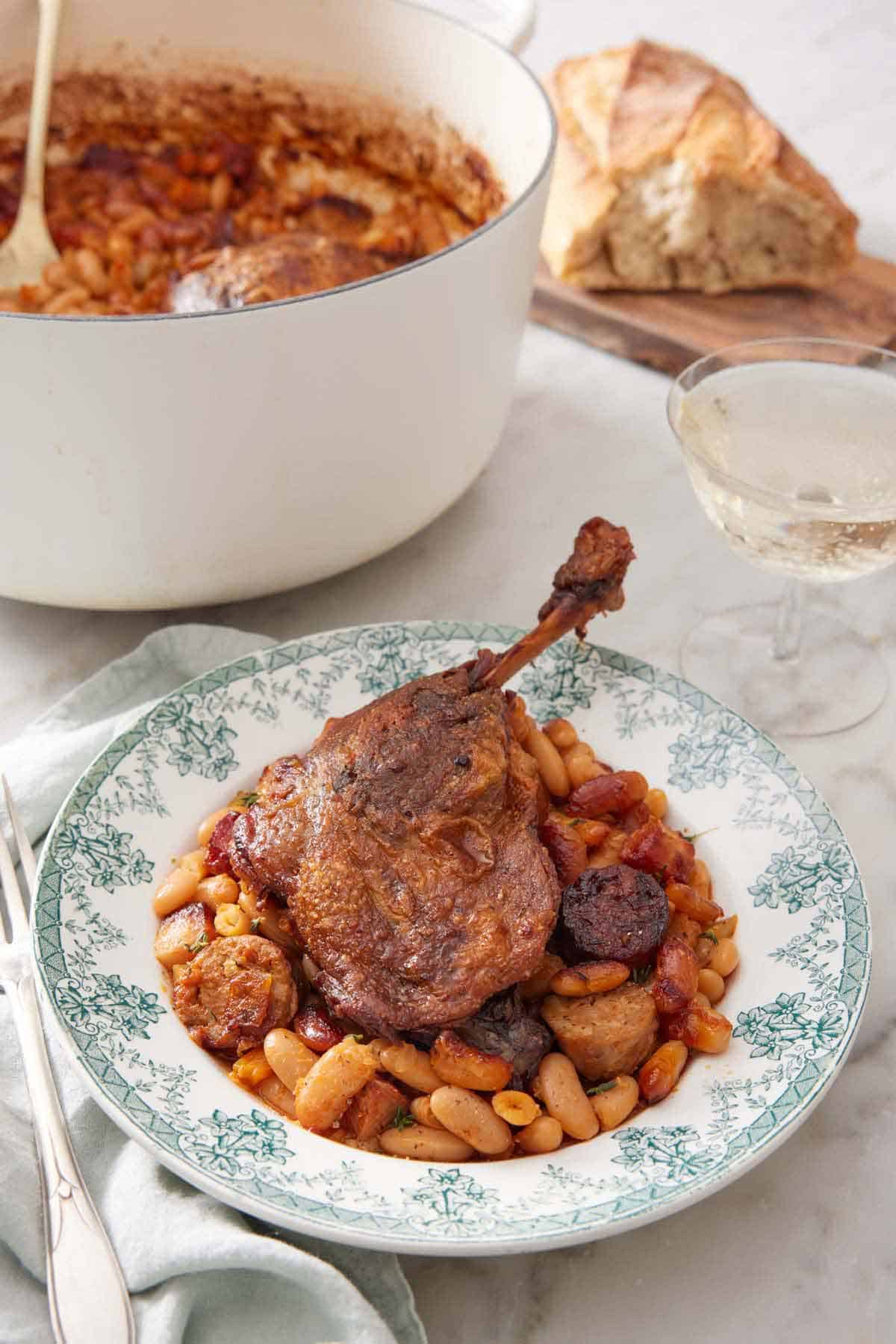 A plate with a serving of cassoulet with a pot with the rest in the background with some wine and bread.
